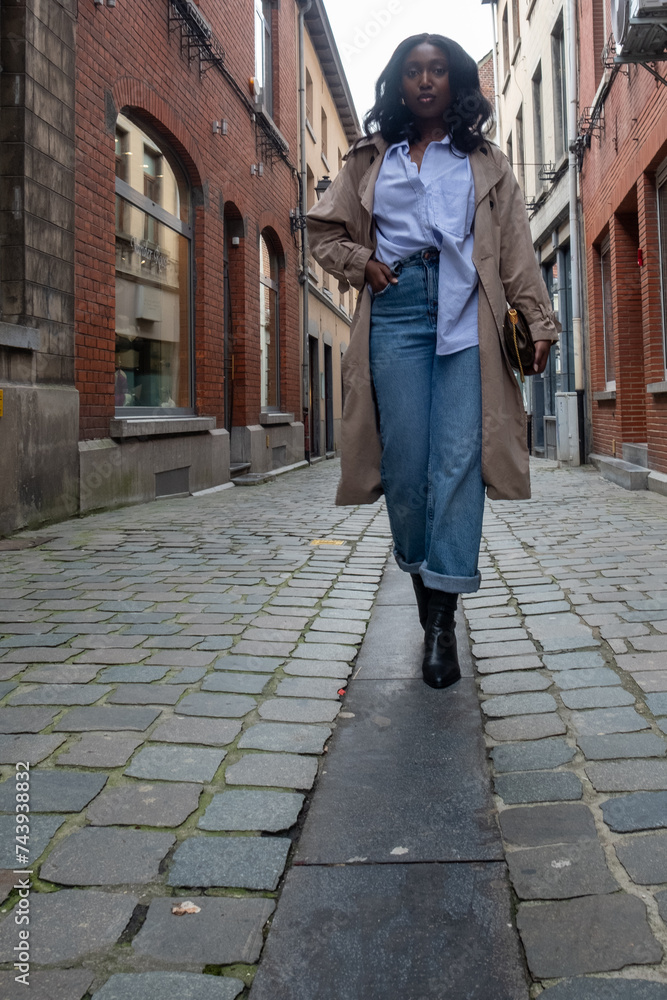 This photograph captures a young African woman walking towards the camera in a cobblestone alleyway, evoking a sense of confidence and independence. Her outfit, consisting of a blue button-down shirt