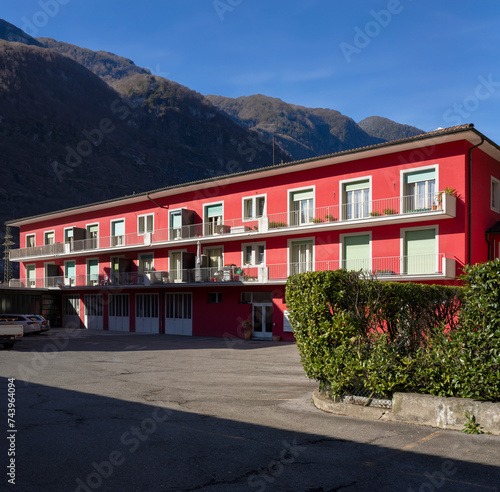 Red railing house with parking area below. Some windows open, others closed by green shutters. Blue sky and mountains behind.