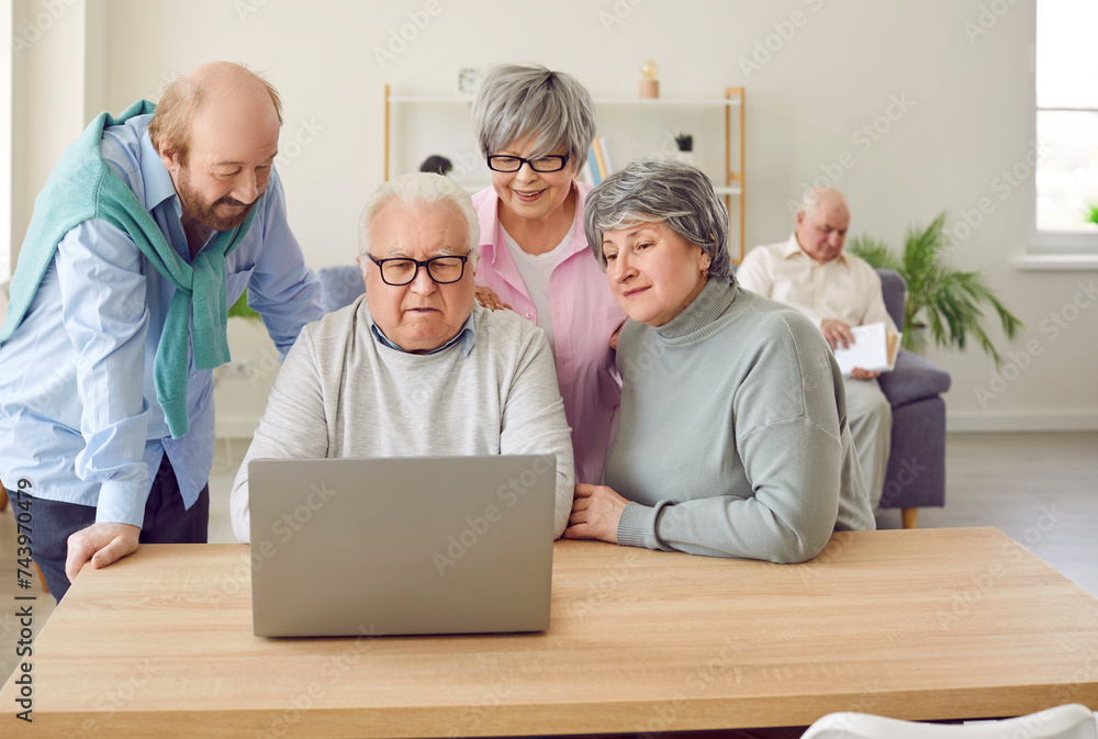 Group of senior people using laptop at retirement home. Several old men and women browsing Internet on notebook computer on wooden table in living room while other man is reading book in background Group of senior people using laptop at retirement home. Several old men and women browsing Internet on notebook computer on wooden table in living room while other man is reading book in background