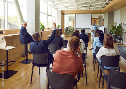 Man raising hand to ask or answer a question on a business meeting. Group of company employees or a team of staff sitting back on chairs and listening to their leader on conference.