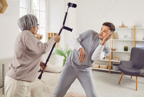 Angry elderly senior woman going to hit young male nurse or doctor with crutch. Enraged exasperated female pensioner standing at home and threatening her scared caretaker with cane.