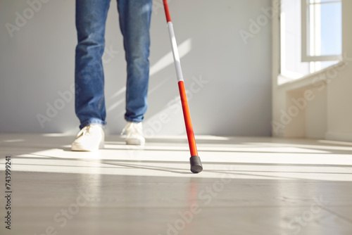 Cropped photo of legs of blind disabled man in casual clothes with long stick walking in empty room at home. Close up person with cane indoors. Blindness, disability and vision problems concept.