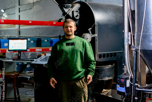 a worker at coffee factory stands near a roasting machine looking at the camera smiling