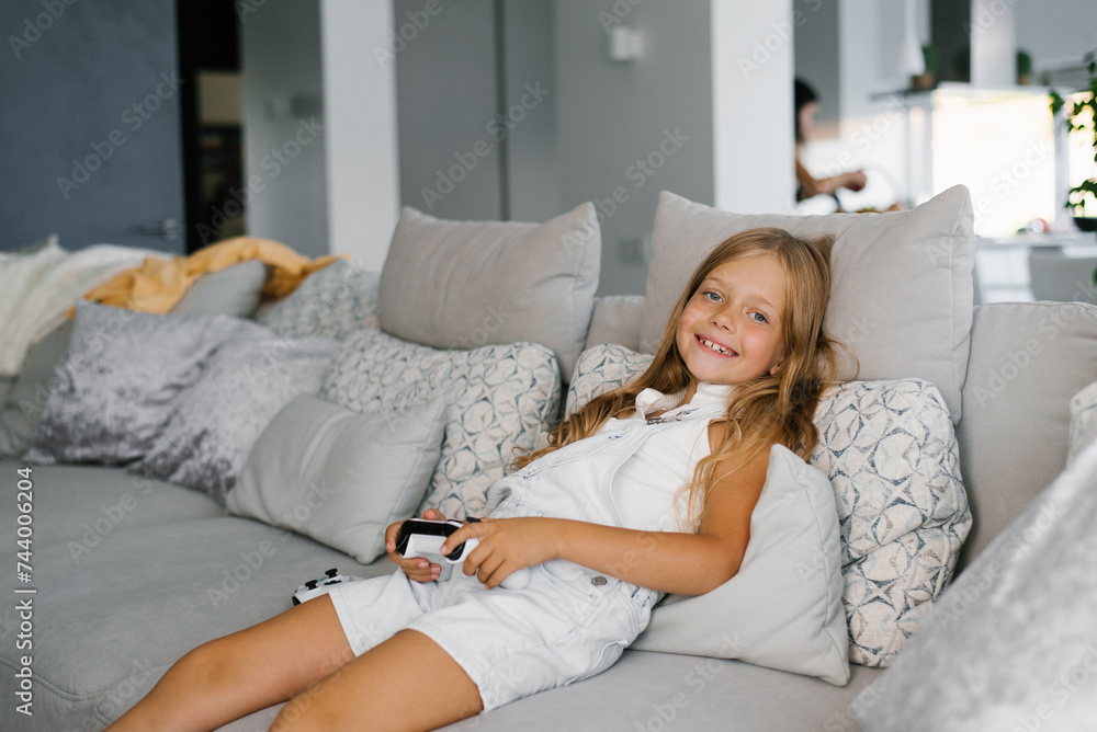 Child girl is smiling, sitting on the couch and holding a game console in her hands Child girl is smiling, sitting on the couch and holding a game console in her hands