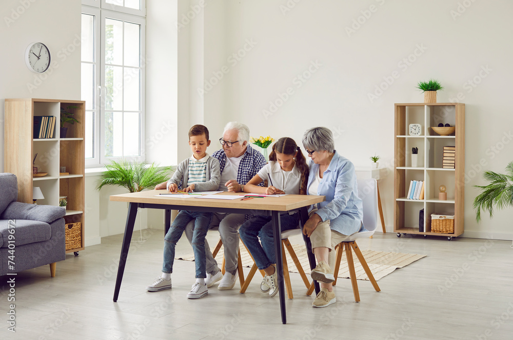 Family spending time together. Grandparents and grandchildren doing quiet indoor activites at home. Grandma, grandpa and children drawing pictures with colored pencils at table in living room interior Family spending time together. Grandparents and grandchildren doing quiet indoor activites at home. Grandma, grandpa and children drawing pictures with colored pencils at table in living room interior