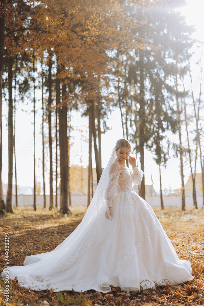 A blonde bride in a white dress with a long train holds the dress and walks down the path covered with autumn leaves. Wedding photo session in nature. Beautiful hair and makeup. Celebration