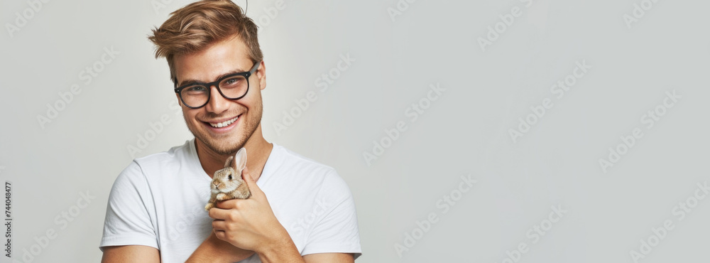 banner a smiling blond man with glasses in a white T-shirt holds an Easter bunny on a light gray background. close-up - obrazy, fototapety, plakaty 