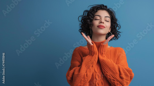 A serene woman in an orange knitted sweater makes an  okay  sign with both hands  eyes closed with a gentle smile  against a vivid blue background.