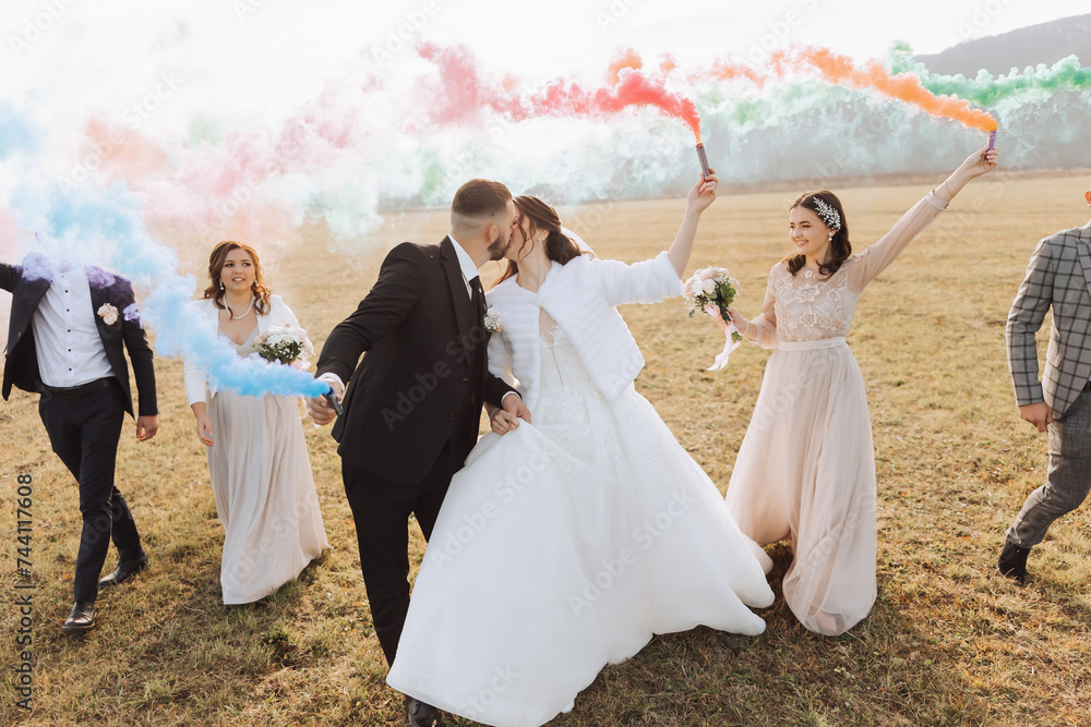 Wedding photo session in nature. Bride and groom and their friends in a field, cheerfully holding colored smoke.