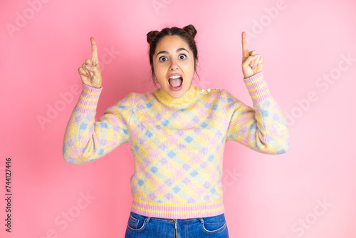 Young beautiful woman wearing casual sweater over isolated yellow background amazed and surprised looking at the camera and pointing up with fingers and raised arms