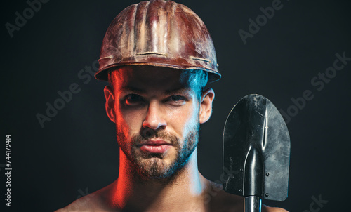 Closeup portrait of construction worker in safety hard hat with spade. Serious bearded man in construction helmet with shovel. Sexy handsome builder, miner, repairman in protective hardhat with spade.