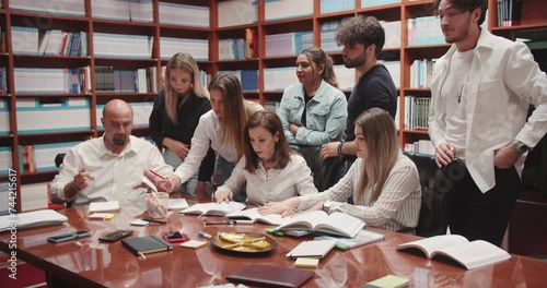 In a bustling university library, a group of diverse college students gathers around a table, engaged in earnest consultation with their professor. This scene embodies the essence of the university photo
