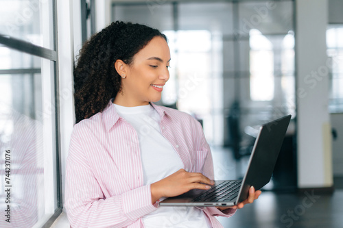 Using gadgets and devices. Pretty, successful brazilian or latino curly haired woman, software manager, company employee, stand in modern office with an open laptop in hands, looks at it, smiles Free Download Using gadgets and devices. Pretty, successful brazilian or latino curly haired woman, software manager, company employee, stand in modern office with an open laptop in hands, looks at it, smiles - GFXLoad