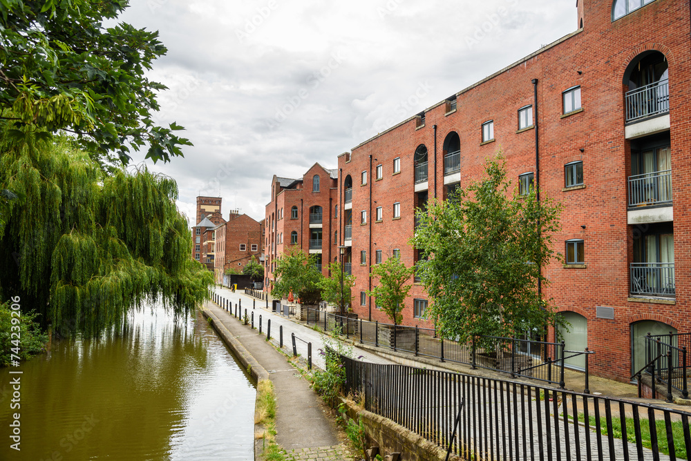 Brick apartment buildings and coverted warehouses along a canal on a cloudy summer day. A cobblestone path lines the canal.