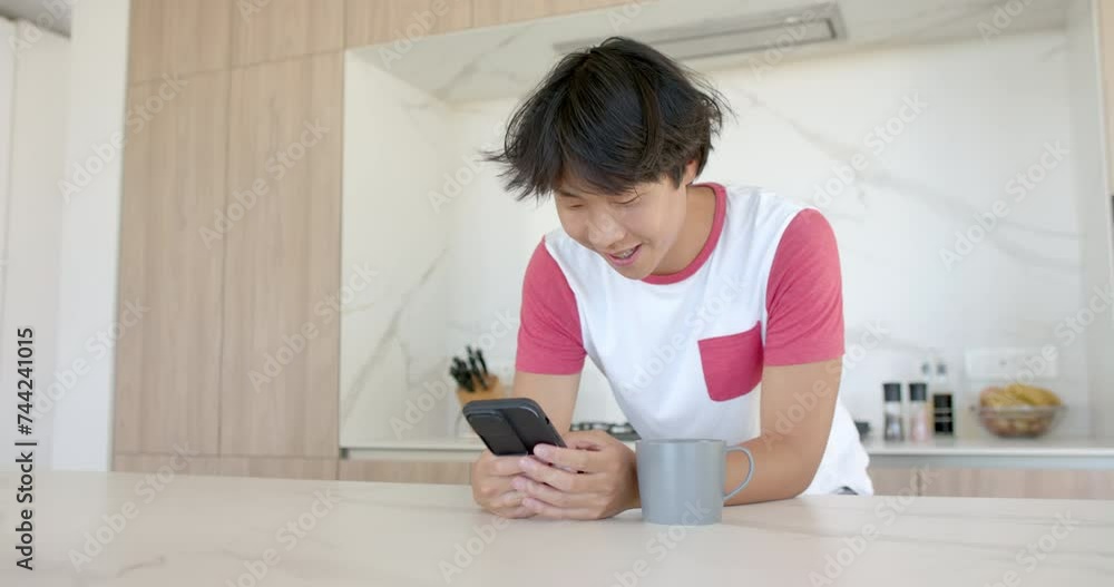 Teenage Asian boy smiles while using his smartphone at home in the kitchen Teenage Asian boy smiles while using his smartphone at home in the kitchen