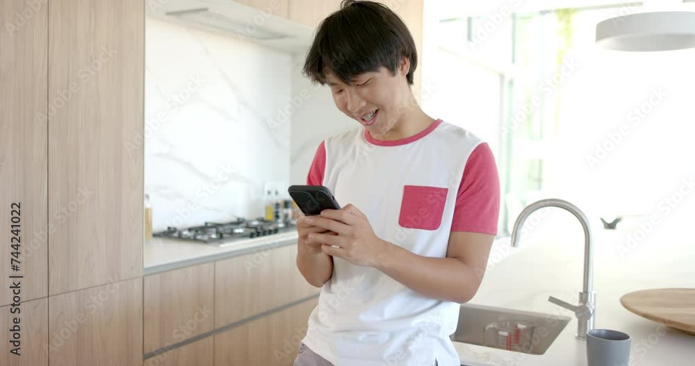 Teenage Asian boy smiles at his phone in a modern kitchen Teenage Asian boy smiles at his phone in a modern kitchen