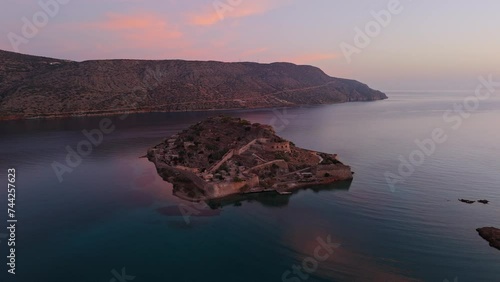 Aerial drone pullback at sunset above Spinalonga island fortress ruins below soft glowing clouds photo