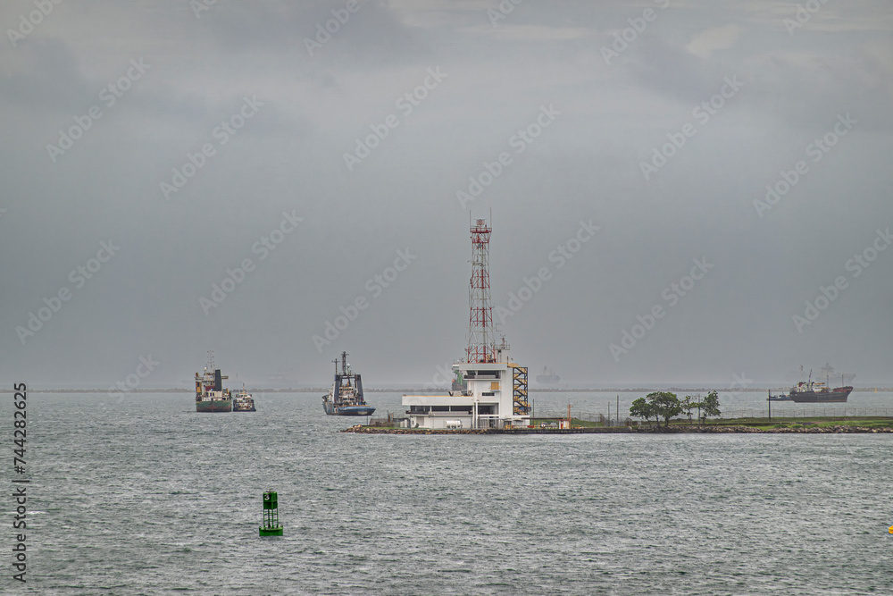 Limon Bay, Colon, Panama - July 24, 2023: Panama Canal Port Captain Office with tall antenna, at end of pier as extension of docks under gray sky. Ships on horizon and green buoy upfront