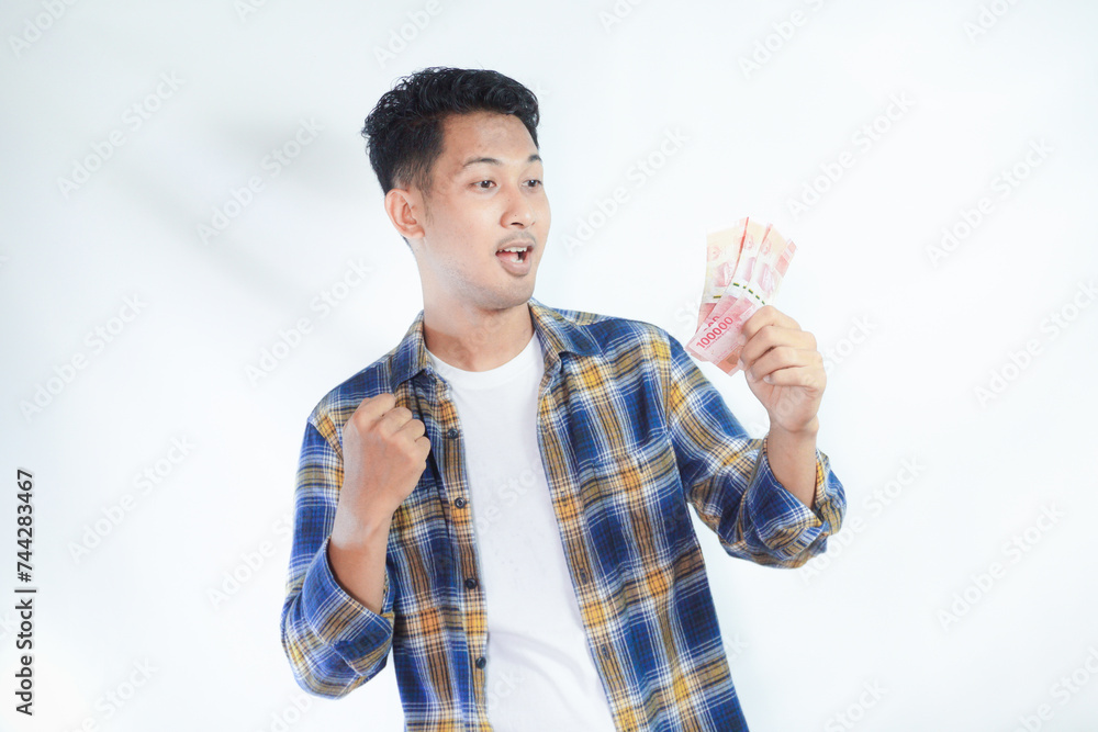 Adult Asian man clenched fist while holding Indonesia paper money and showing excited expression Adult Asian man clenched fist while holding Indonesia paper money and showing excited expression