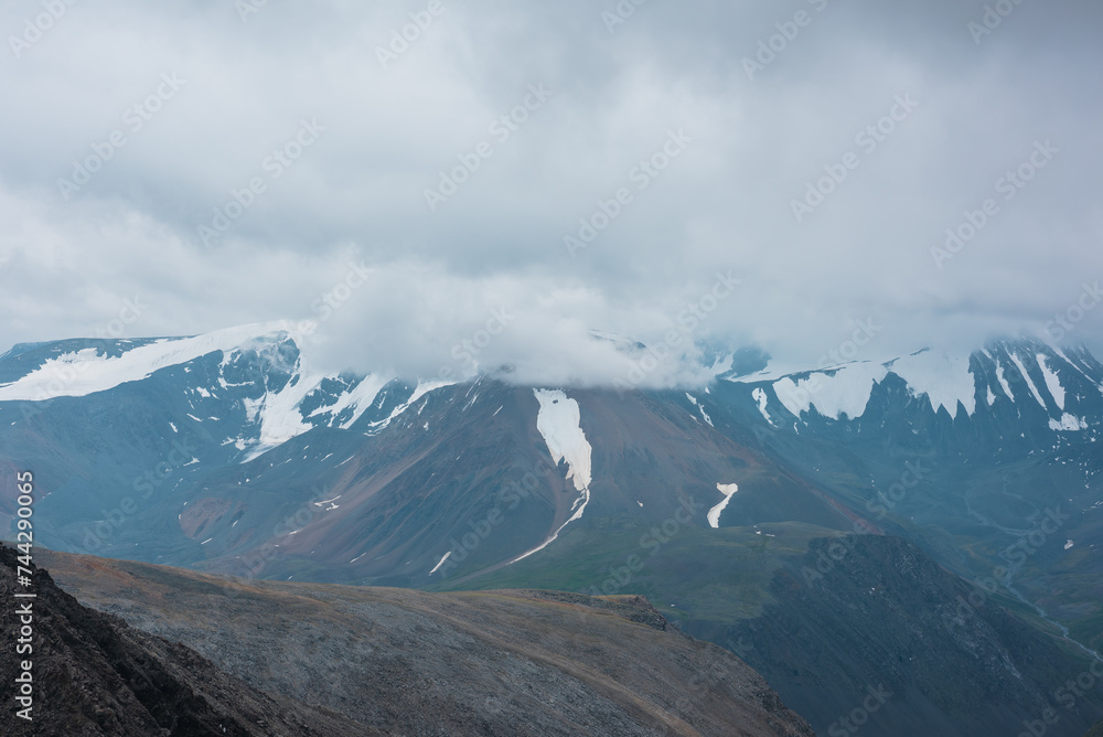 Large snow mountain range silhouette in rainy low clouds under gray sky. Stony hill and rocky pass in far away in bad weather. Low cloud on big snowy mountains in light transparent mist in overcast.