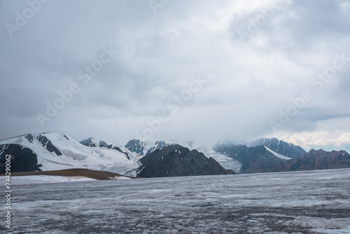 Dramatic vast misty top view from ice pass to big glacier tongue among sharp rocks and large snow-capped mountains in rainy low clouds. Dark atmospheric mountain silhouettes in rain in gray cloudy sky
