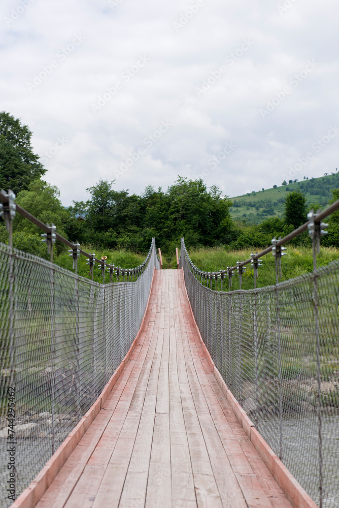 Obraz premium A new wooden bridge over a mountain river on cables. Ukrainian Carpathian Mountains. A bridge over a mountain river. Green grass and trees.