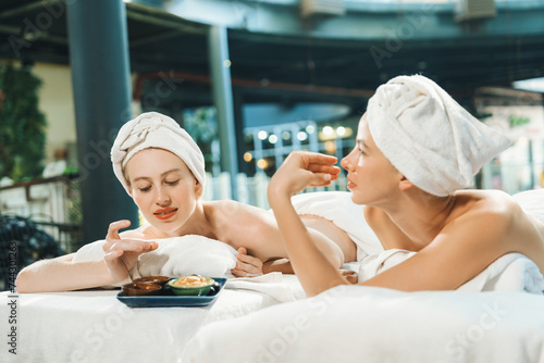 Couple of beautiful young girls lie on spa bed during interested in homemade beauty facial mask. Attractive woman touching herbal facial mask. Surrounded with nature environment. Tranquility.