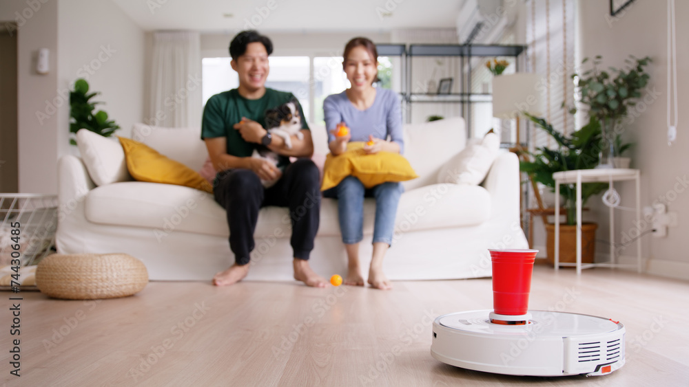 Internet of things smart urban house IOT appliances better life. Asia people man woman joy win fun beer ping pong ball throw cup game to robot hoover clean floor at home sofa living room with pet dog.