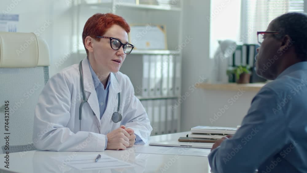Caucasian female physician wearing white coat and stethoscope over neck consulting African American patient during medical appointment in clinic Caucasian female physician wearing white coat and stethoscope over neck consulting African American patient during medical appointment in clinic