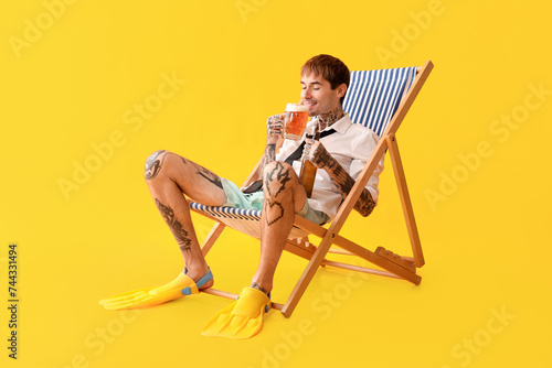 Tattooed young man with bottle and mug of beer sitting on beach chair against yellow background
