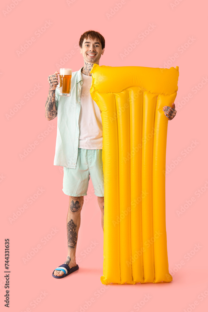 Obraz premium Tattooed young happy man with inflatable mattress and mug of beer on pink background