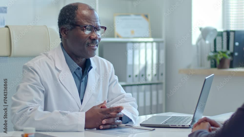 Senior Black doctor sitting at desk, asking questions and listening attentively to female patient during appointment in medical office Senior Black doctor sitting at desk, asking questions and listening attentively to female patient during appointment in medical office