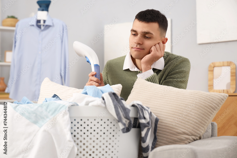 Obraz premium Tired young man with steamer and basket full of laundry at home