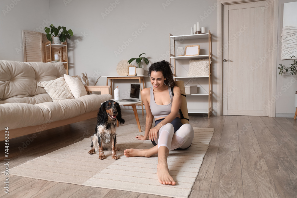Sporty young African-American woman with yoga mat and cocker spaniel sitting on floor at home