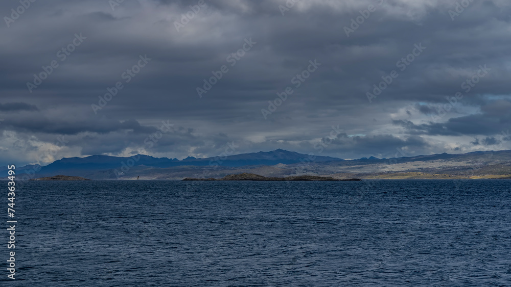 The coastline of the Tierra del Fuego archipelago is visible from the Beagle Channel. The Andes mountains against a cloudy sky. Ripples on the blue water of the strait. Argentina. The coastline of the Tierra del Fuego archipelago is visible from the Beagle Channel. The Andes mountains against a cloudy sky. Ripples on the blue water of the strait. Argentina.