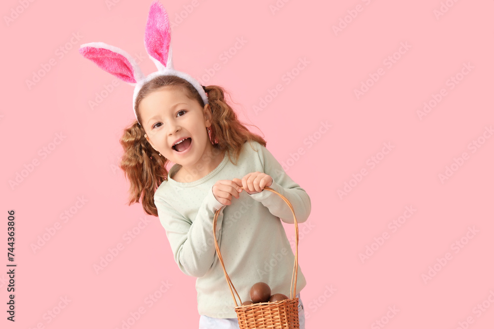 Cute little girl in bunny ears with basket of chocolate Easter eggs on pink background
