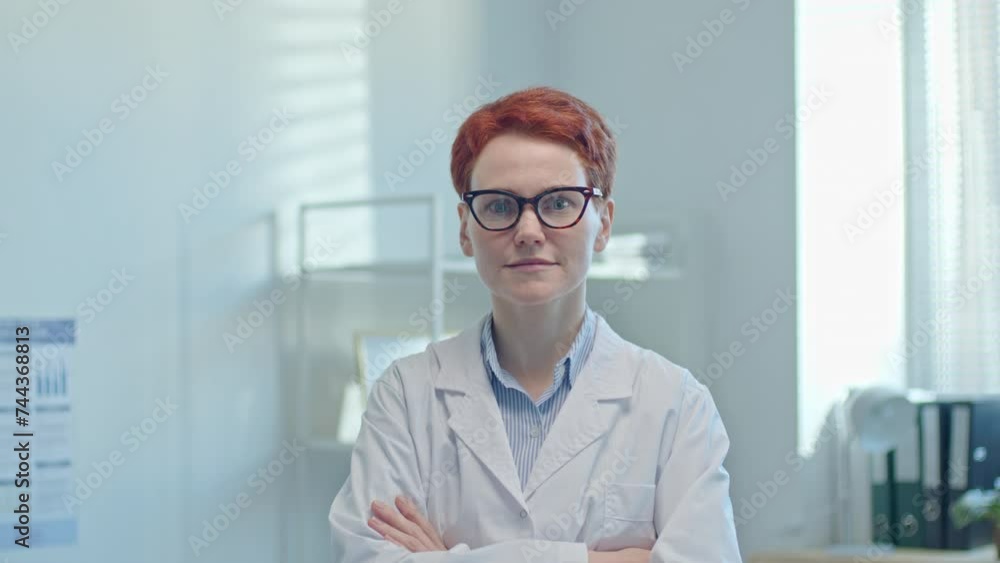 Redhead female doctor in white coat posing for camera with arms crossed at work in medical office. Video portrait