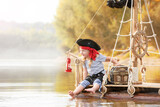 Child in a pirate costume plays on a wooden raft at sunset. Girl pretends to be the captain of a ship with black sails and a flag. Funny kid dreams of adventure and travel.