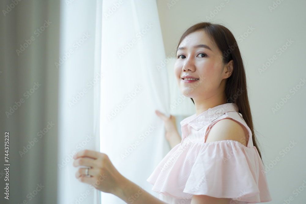 Happy Beautiful young Asian woman opening window curtain in the morning. Portrait of Pretty Asian young woman looking the sun rise outside through the window peacefully, Asian girl portrait close up. Happy Beautiful young Asian woman opening window curtain in the morning. Portrait of Pretty Asian young woman looking the sun rise outside through the window peacefully, Asian girl portrait close up.