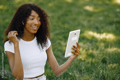 Female african student sitting on a grass and using a tablet fo a video call