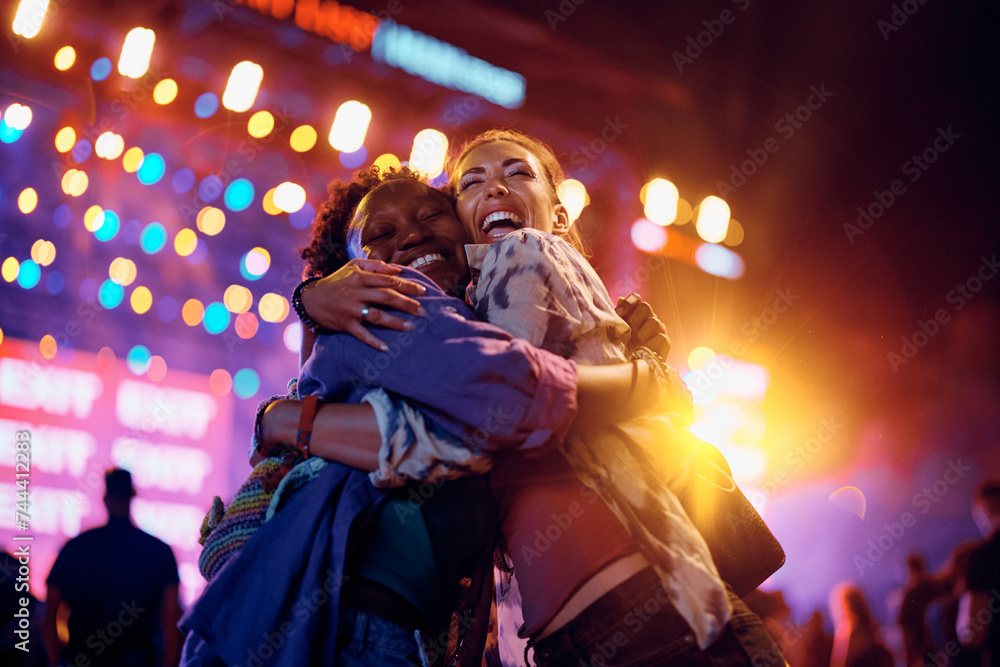 Cheerful female friends embracing in front of music stage during summer festival at night.