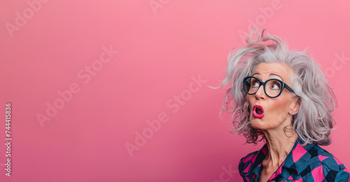 surprised mature woman with glasses and voluminous hairstyle on a pink background. face of surprise and curiosity. pink background of tenderness and femininity. copy space for advertising or banners