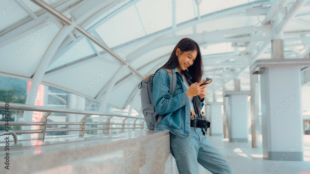 Young asian woman smiling using mobile smart phone outdoor. Happy female tourist wearing jeans jacket and holding smartphone at public