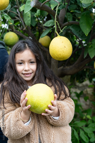 Young girl with a beaming smile holding a large pomelo, surrounded by green foliage.