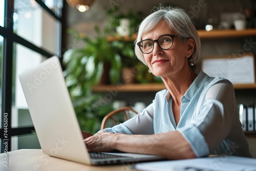 Smiling mature middle aged businesswoman using laptop working on sitting at desk. Happy old businesswoman hr holding cv interviewing distance applicant, senior seeker searching job, Generative AI