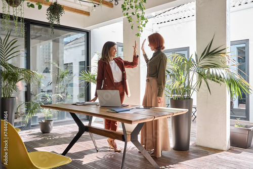 Two smiling female employees or entrepreneurs, happy professional business women celebrating work success and partnership support together giving high five standing in green office. Authentic photo. photo