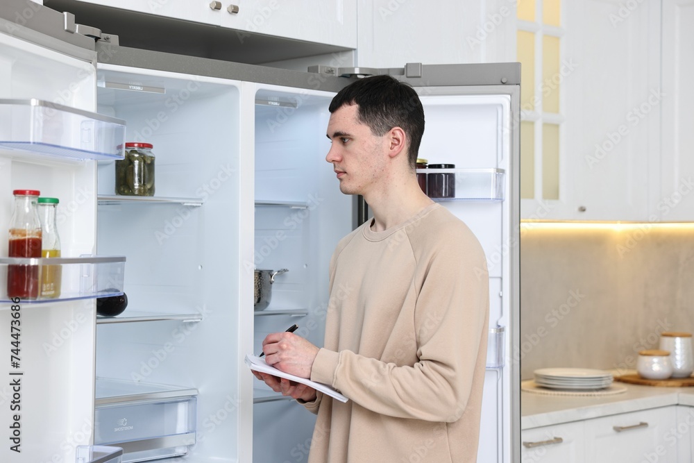 Man writing notes near empty refrigerator in kitchen