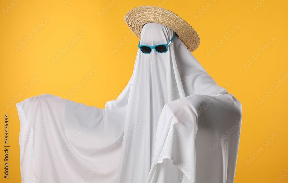 Person in ghost costume, sunglasses and straw hat on yellow background Person in ghost costume, sunglasses and straw hat on yellow background