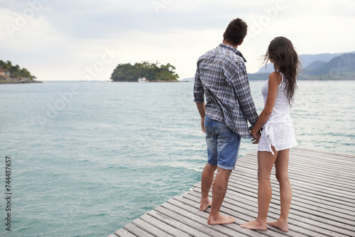 Couple, jetty and holding hands by ocean on vacation, security and relax by water on holiday. People, travel and bonding for relationship in outdoor, support and back on weekend trip to sea or nature