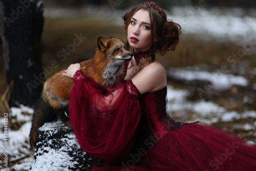 Beautiful young woman in long red dress sitting near fox in the forest. Fantasy fairytale portrait of red haired girl